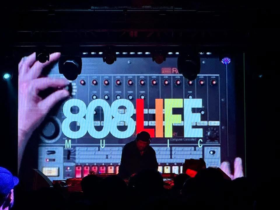 DJ silhouette in front of the 808 Life screen and drum machine visuals